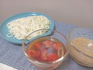 ランチマットの上に置かれためんつゆとトマト、すりゴマ味噌のつけ汁とそうめんの写真